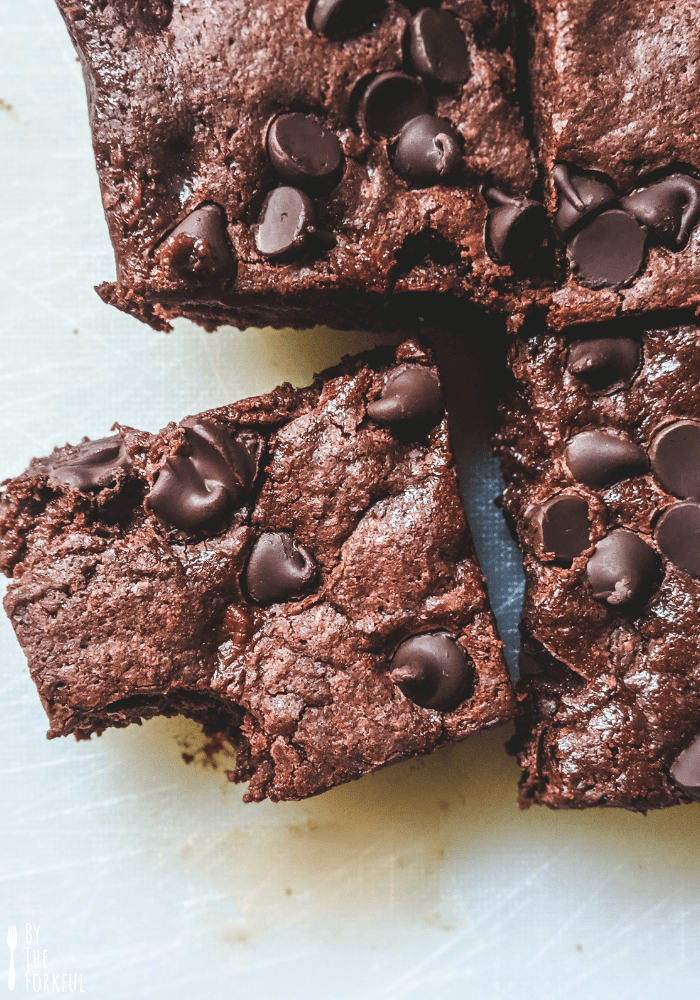 Vegan chocolate brownies topped with chocolate chips on a white chopping board. One brownie is tilted at an angle and has a bite taken out of it.
