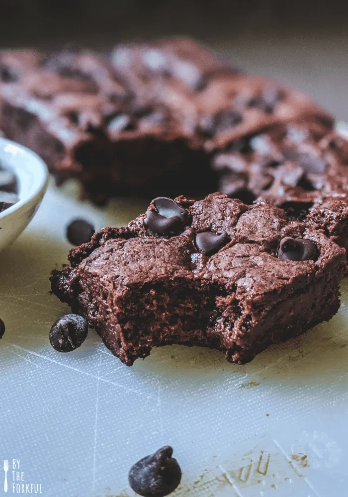 Vegan brownies in the background on a white chopping board. A small white bowl of chocolate chips can just be seen, and a brownie is in the foreground with a bite taken out of it.