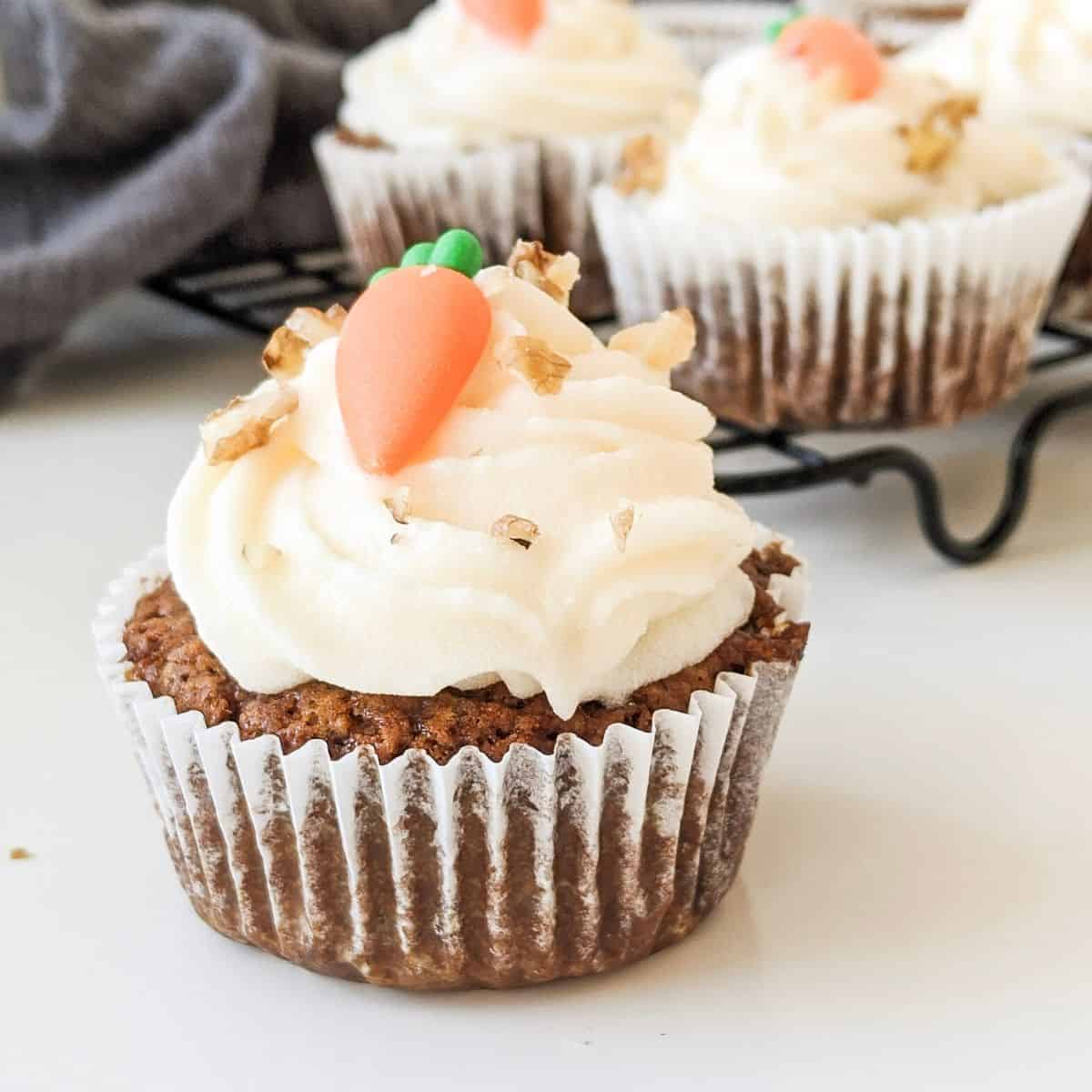 Vegan carrot cake cupcake topped with cream cheese frosting and a sugar carrot decoration. More cakes are in the background.