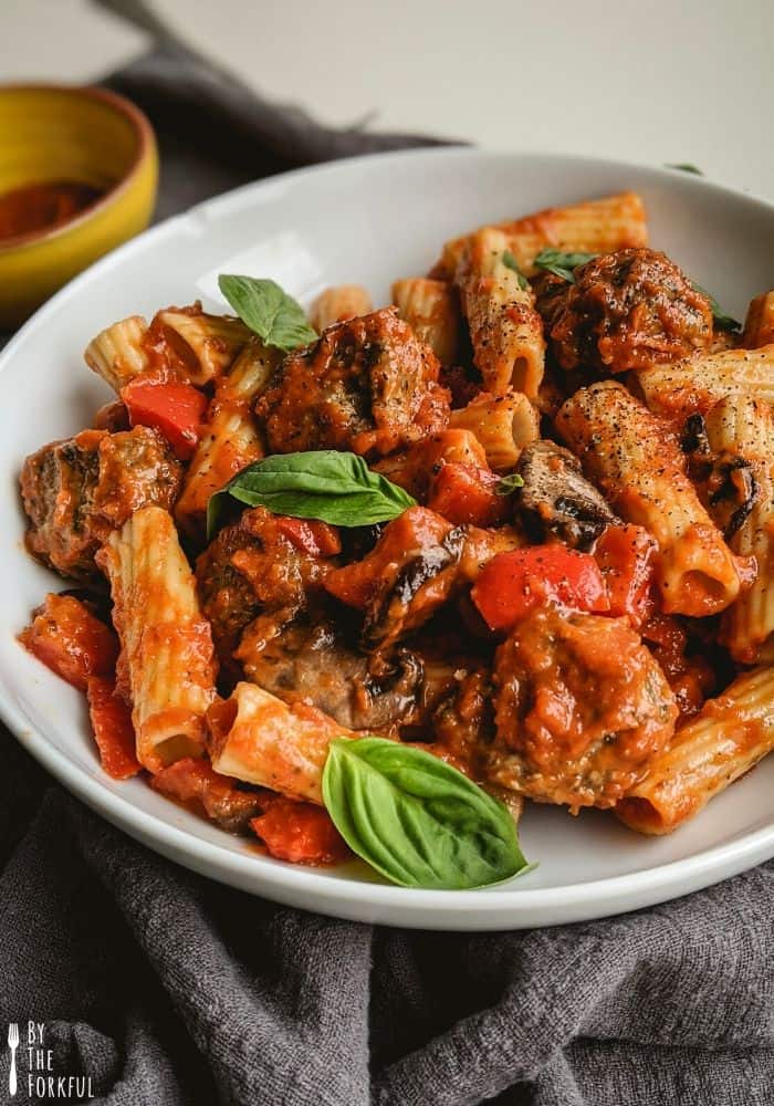 Vegan meatball pasta with tomato sauce, rigatoni pasta, roasted pepper and mushrooms, and topped with fresh basil in a white bowl.