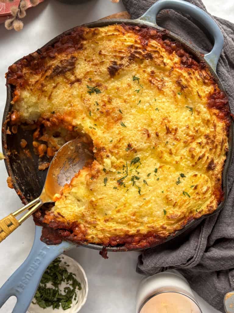 a overhead shot of vegetarian shepherd's pie with golden crispy mashed potatoes cooked in a cast iron skillet. A serving spoon has taken a scoop from the pan