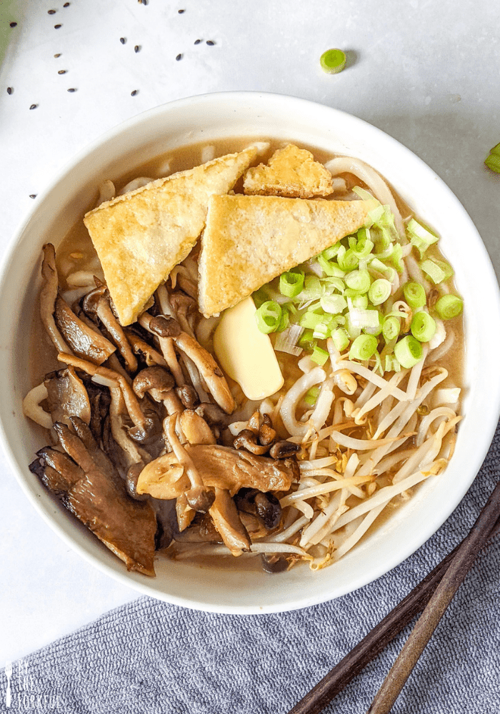 Bowl of udon noodles in a mushroom broth, topped with mixed mushrooms, cripsy tofu cut into triangles, sping onions, and bean sprouts.