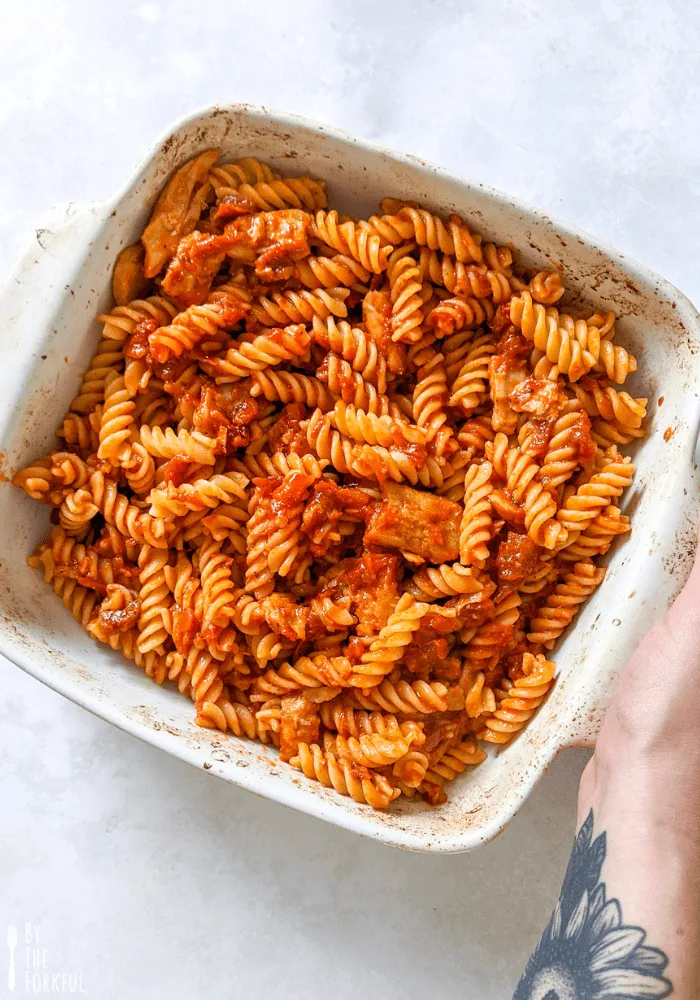 Vegan chicken and tomato pasta in a baking dish. A hand holds the side of the dish.