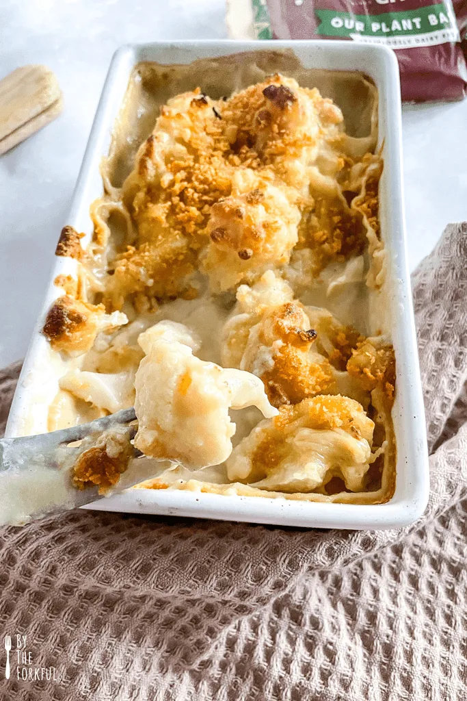 A delicious vegan cauliflower cheese with a fork just taking a bite out of the white baking dish.