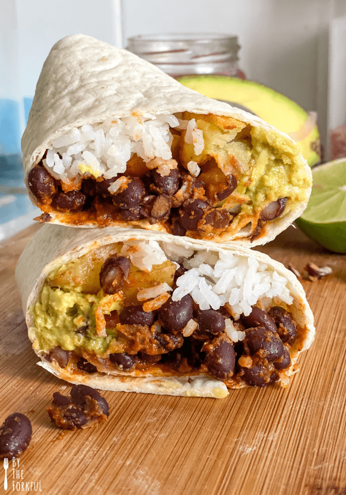 A black bean burrito with rice and avocado. The burrito is cut in half with one half stacked on top of the other on a wooden cutting board.