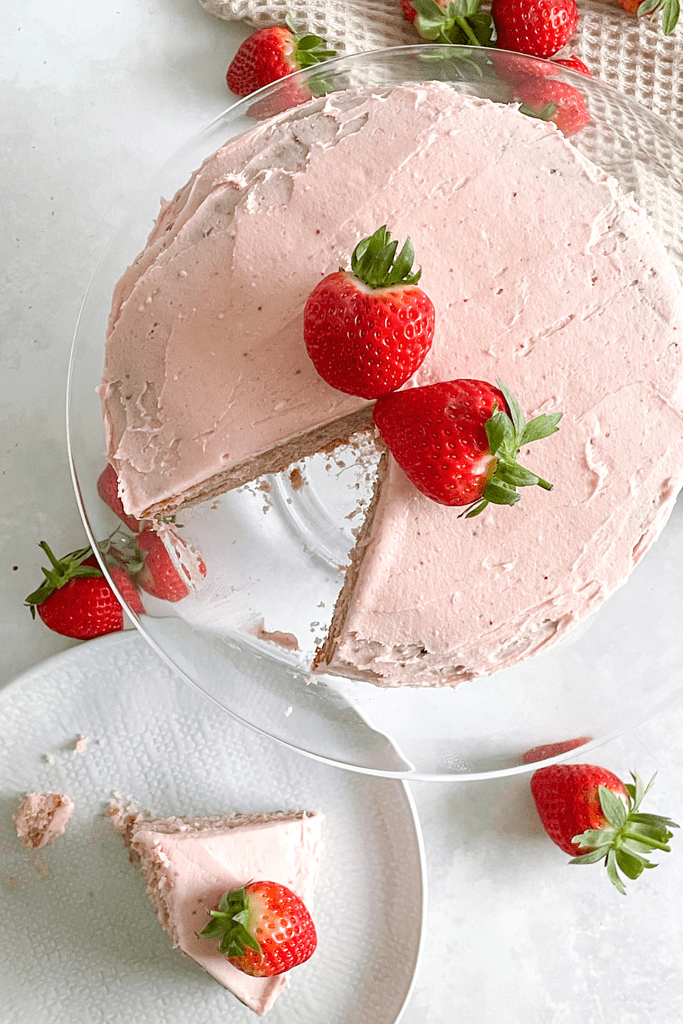 A birdseye view of a pink vegan strawberry cake topped with bright red strawberries. A slice has been cut out of the cake and sits on a plate to one side.