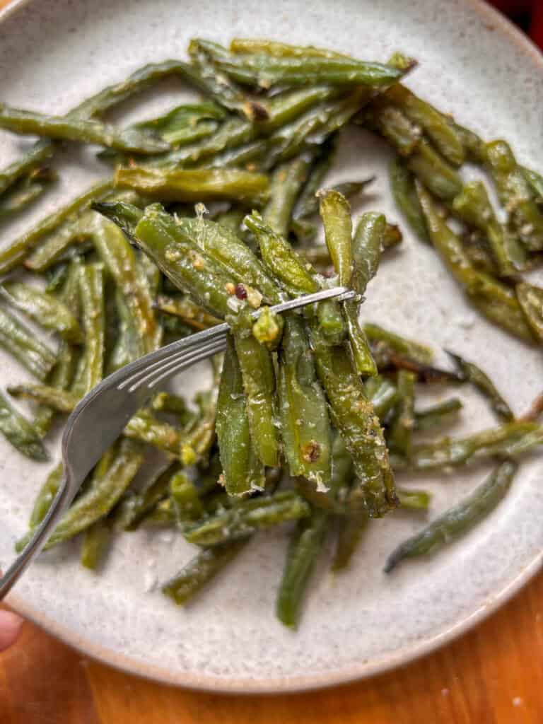 a plate of roasted frozen green beans, with a fork held above it with a large portion of the green beans speared.