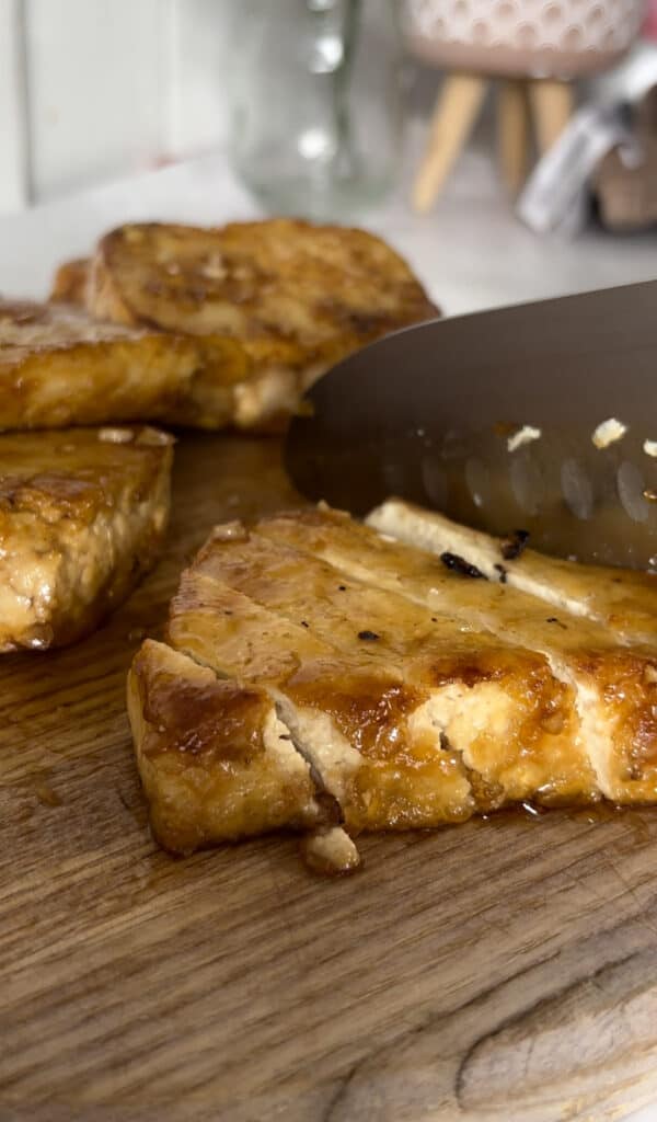 A slice of honey garlic tofu being sliced into thin strips on a cutting board.