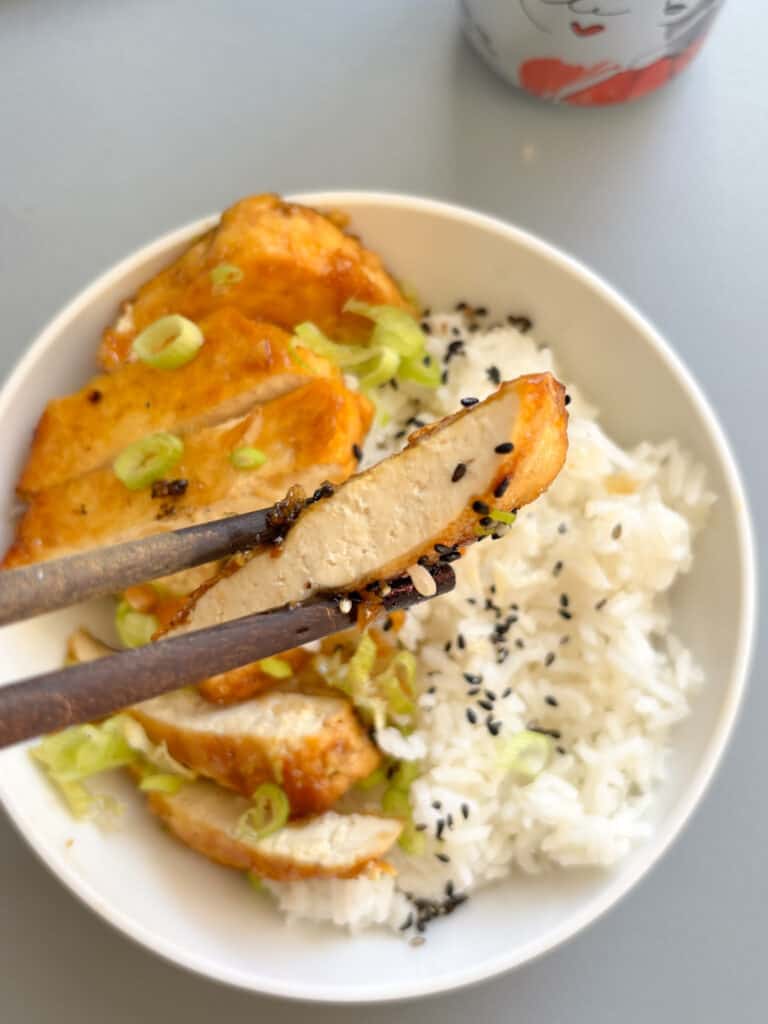 a close up shot of a slice of honey garlic tofu, with the remaining bowl (paired with white rice) and on a blue surface in the back ground.