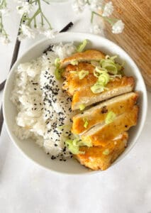 a white bowl with white rice and crispy honey garlic tofu topped with sliced spring onion and black sesame seeds.