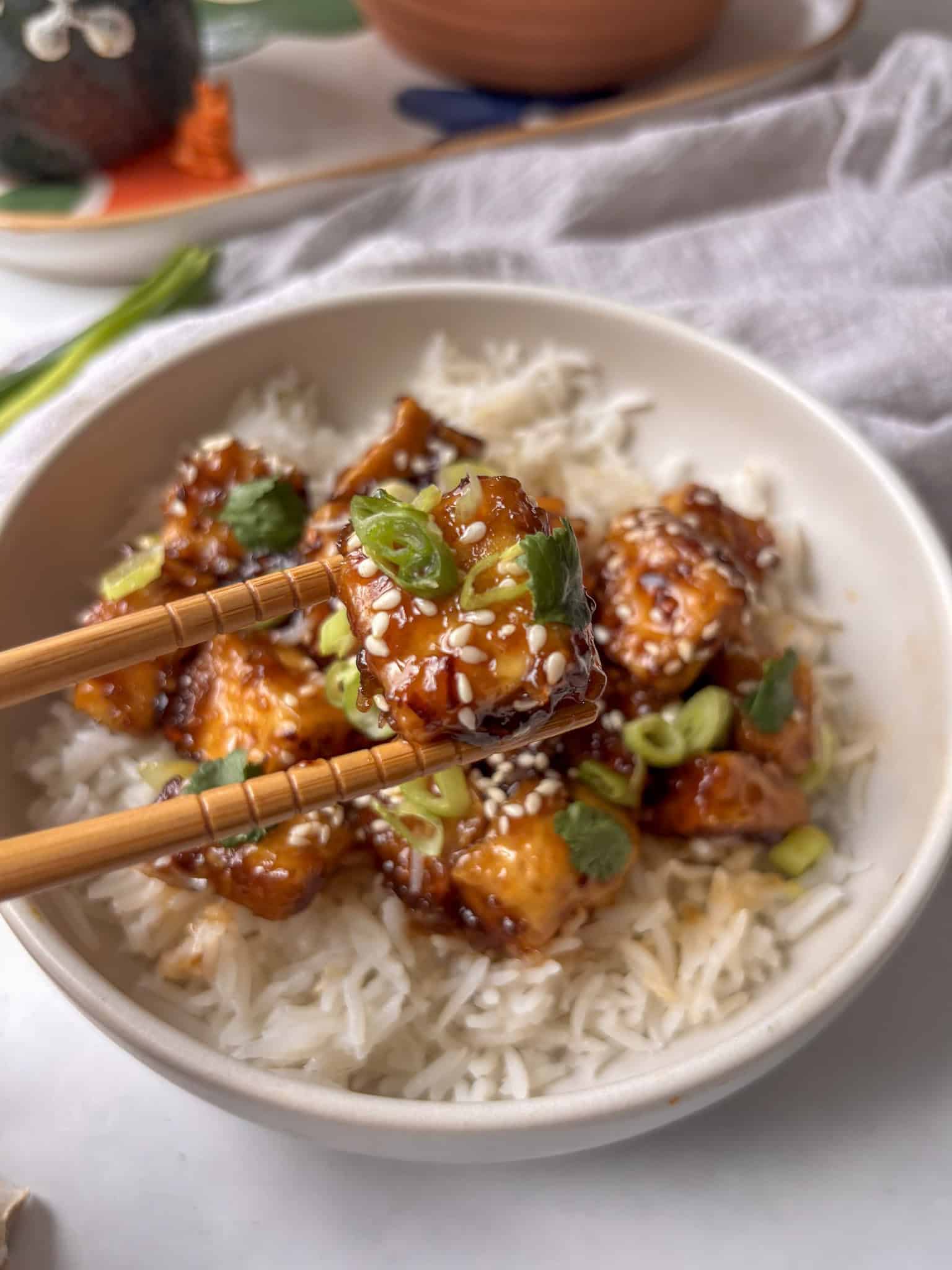 honey garlic tofu on top of rice, with scallions and sesame seeds on top in the background, with a cube of honey garlic tofu being held in chopsticks in the foreground
