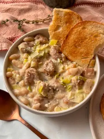 creamy sausage, leek, and white bean stew topped with Pecorino cheese and cracked black pepper in a white bowl, with a spoon on the side.