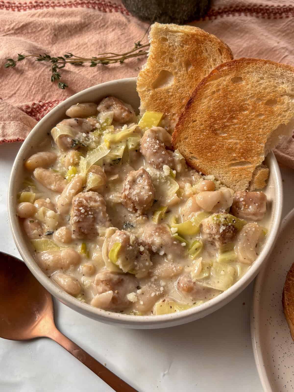 creamy sausage, leek, and white bean stew topped with Pecorino cheese and cracked black pepper in a white bowl, with a spoon on the side.