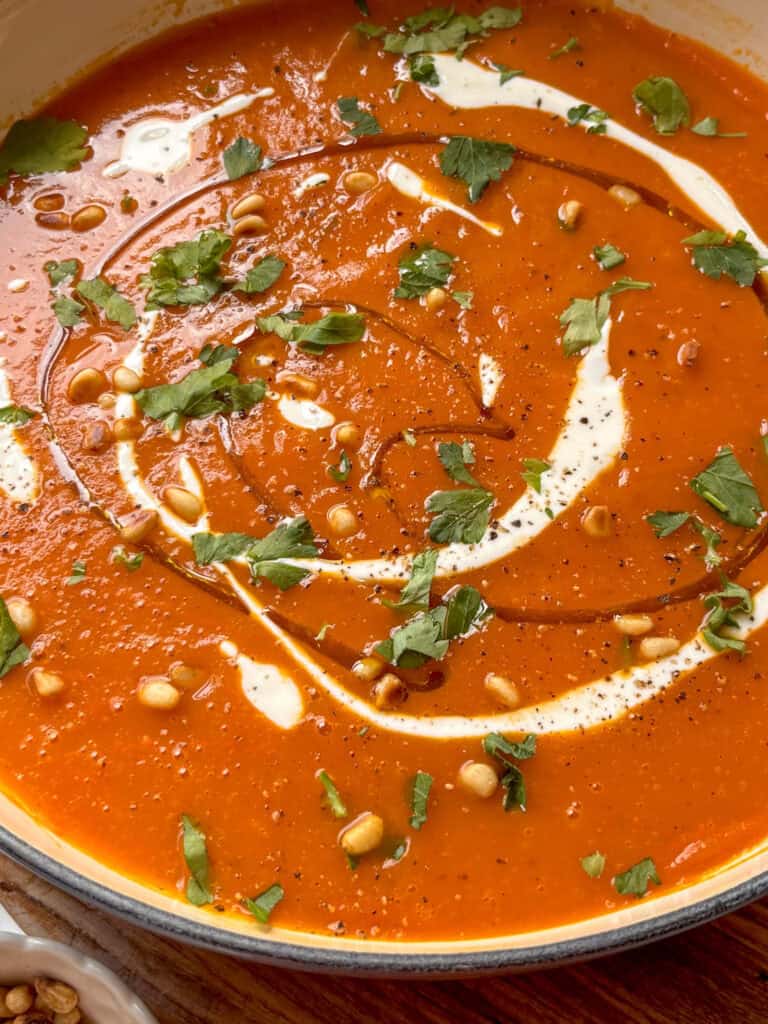 a close up shot of a vibrant butternut squash and red pepper soup drizzled with olive oil, cream, parsley, and pine nuts