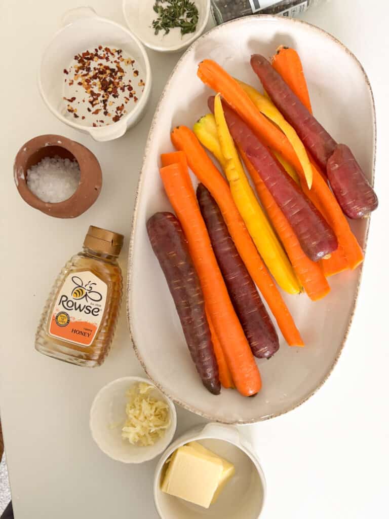 An overhead shot of the ingredients require for this honey butter carrots recipe: rainbow carrots, honey, garlic, butter, chili, thyme, salt, and pepper