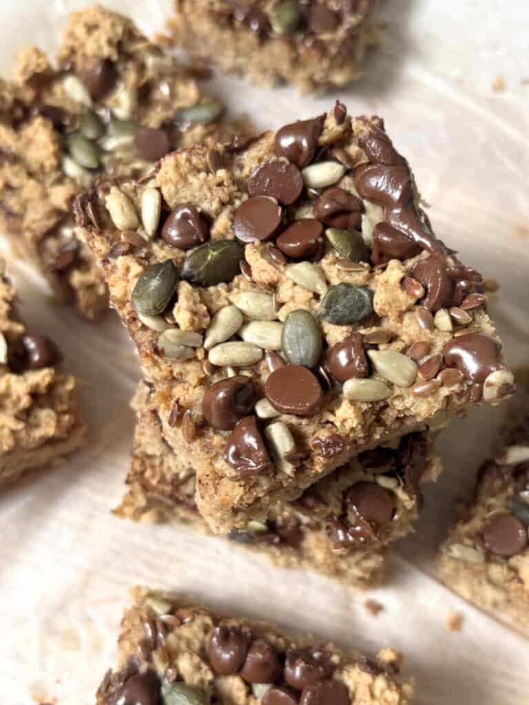 a protein flapjack bar stacked on top of others. the top of the flapjack is decorated with dark chocolate chips and a mix of sunflower seeds, sesame seeds, and lindseeds.