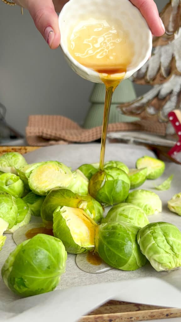 halved brusssel sprouts on a baking tray lined with baking paper. They are being drizzled with a mixture of maple syrup and olive oil 