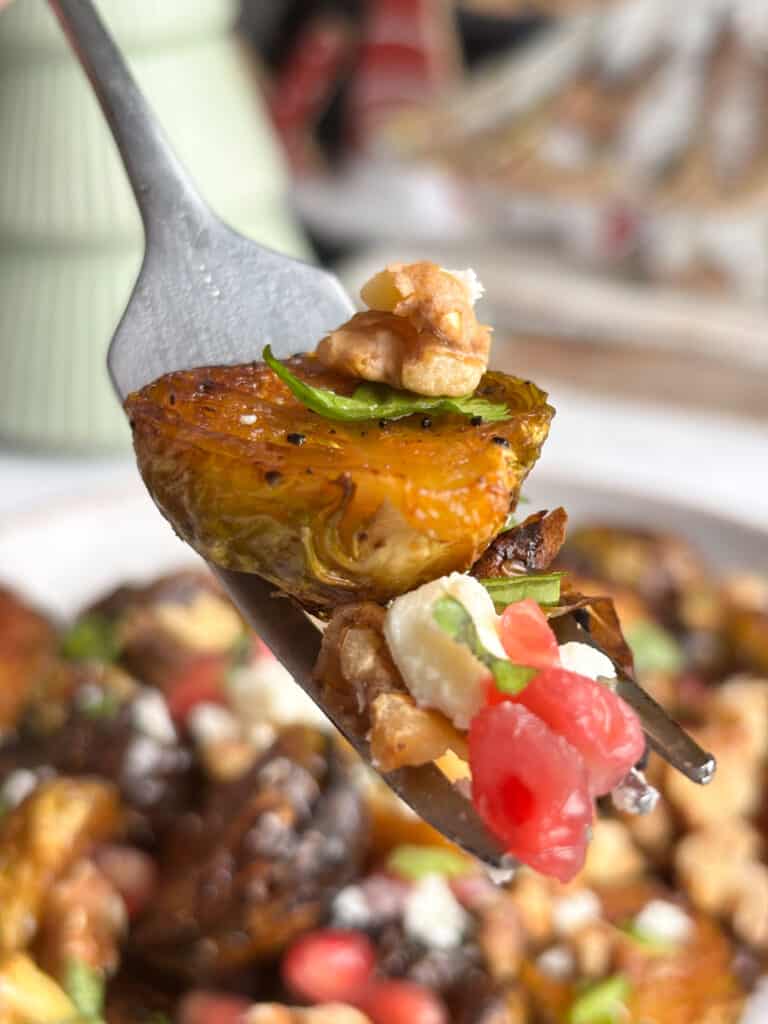 A close up shot of a fork with a balsamic sprout, chopped walnut, pomegranate, feta and parsley on it. The serving plate of balsamic sprouts can be seen in the background
