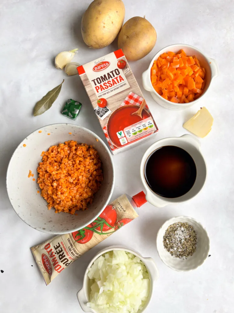 An overhead shot of the ingredients you need to make this vegan lentil hotpot: diced onions, diced carrots, chopped tomatoes, red lentils, tomato puree, tamari, seasonings, potatoes, vegetable stock, and garlic