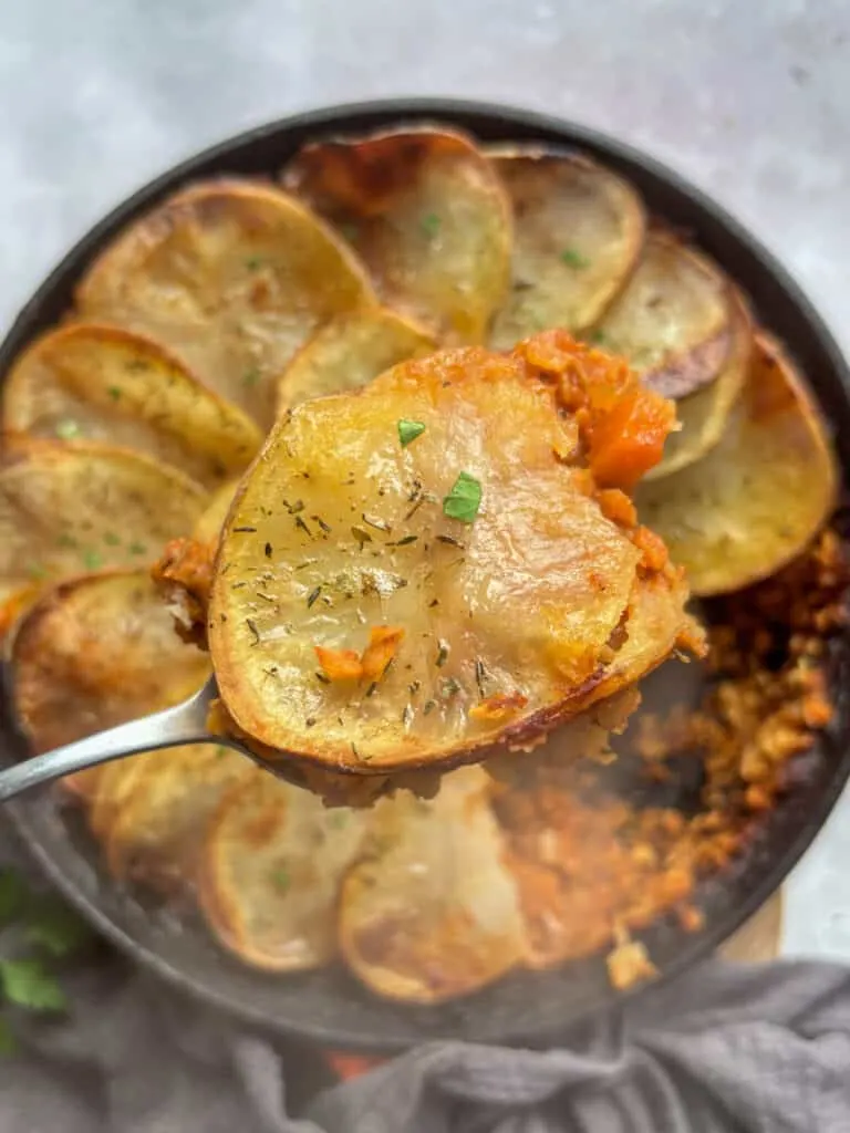 a spoonful of vegan lentil hotpot with a slice of crispy potato on top. The full skillet of vegan hotpot is underneath slightly burred out.