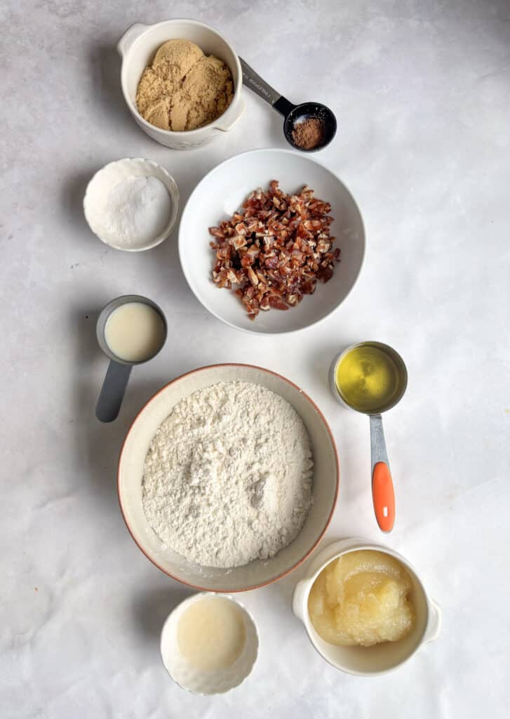 an overhead shot of all the ingredients needed for vegan applesauce cake: plain flour, almond milk, apple sauce, apple cider vinegar, olive oil or coconut oil, dates, baking powder, baking soda, cinnamon, coconut sugar or light brown sugar