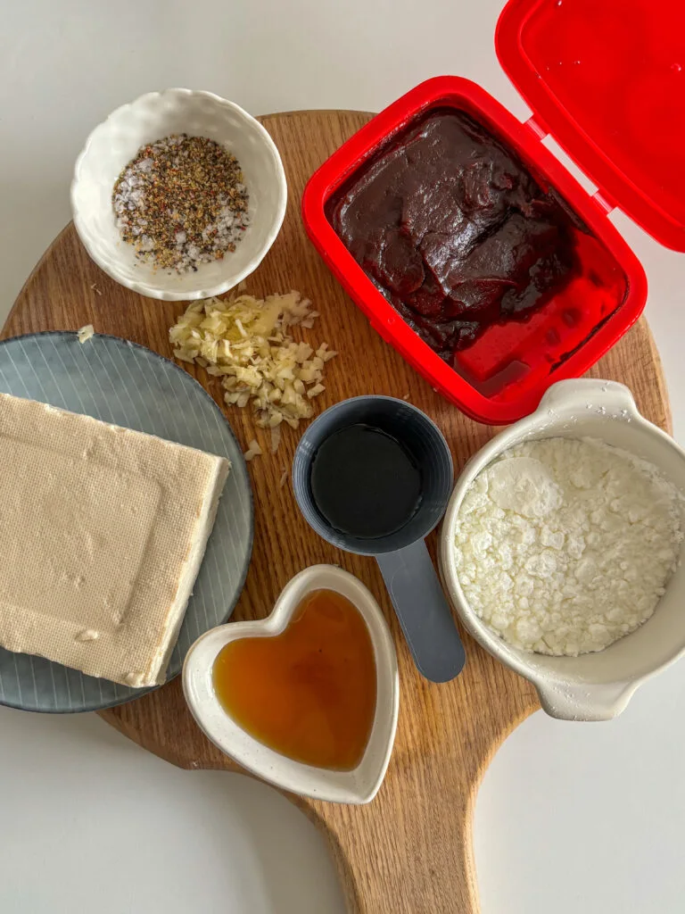 an overhead shot of all the ingredients needed to make gochujang tofu: gochujang (red chilli paste), extra-firm tofu, minced garlic, soy sauce, maple syrup, corn flour, water, and seasonings.