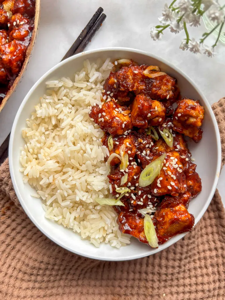 a white bowl of rice and spicy gochujang tofu topped with sesame seeds and sliced scallions
