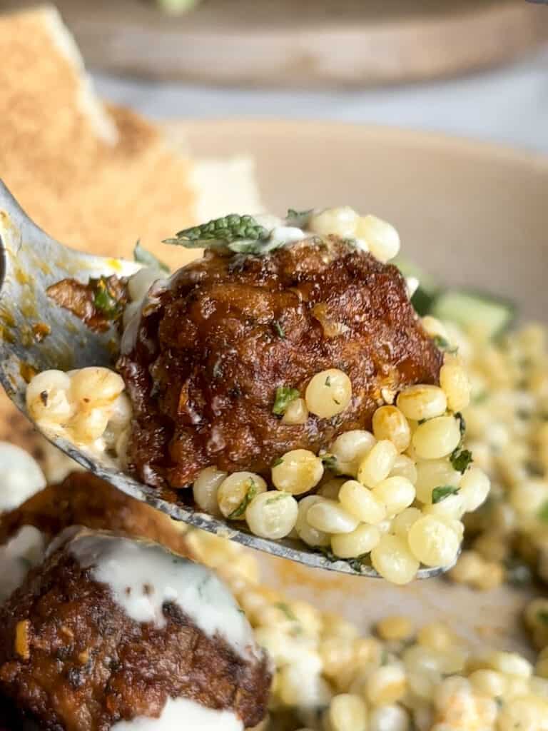 a close up shot of a harissa meatball with pearl couscous and herbs on a spoon. The rest of the dish can be seen in the background.