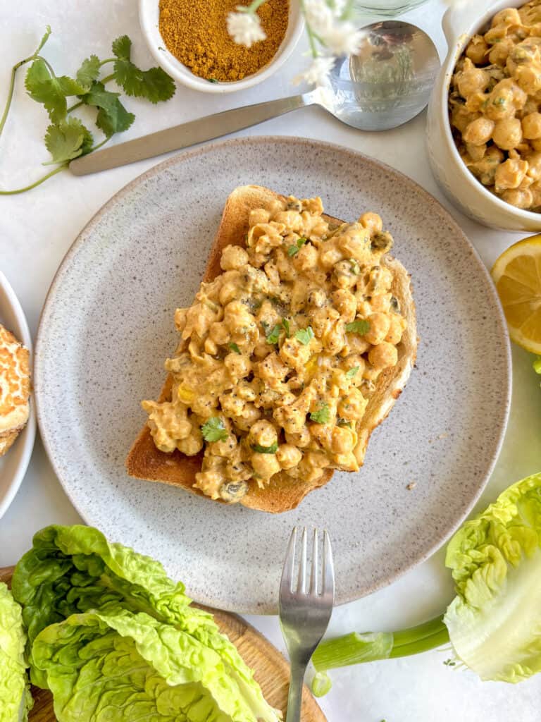 a plate with a slice of toasted sourdough toast loaded with coronation chickpeas. The plate is decoratively surrounded by a small bowl of curry powder, lemons, leftover coronation chickpeas, and some coriander sprigs.