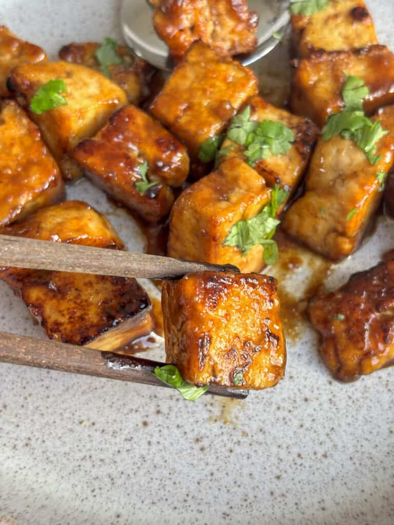 a cube of crispy marinated tofu being held by chopsticks, with the remaining dish in the background