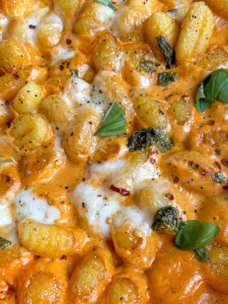 a close up shot of the crispy, golden gnocchi in vodka sauce topped with melted mozzarella and fresh basil leaves, and a scattering of black pepper