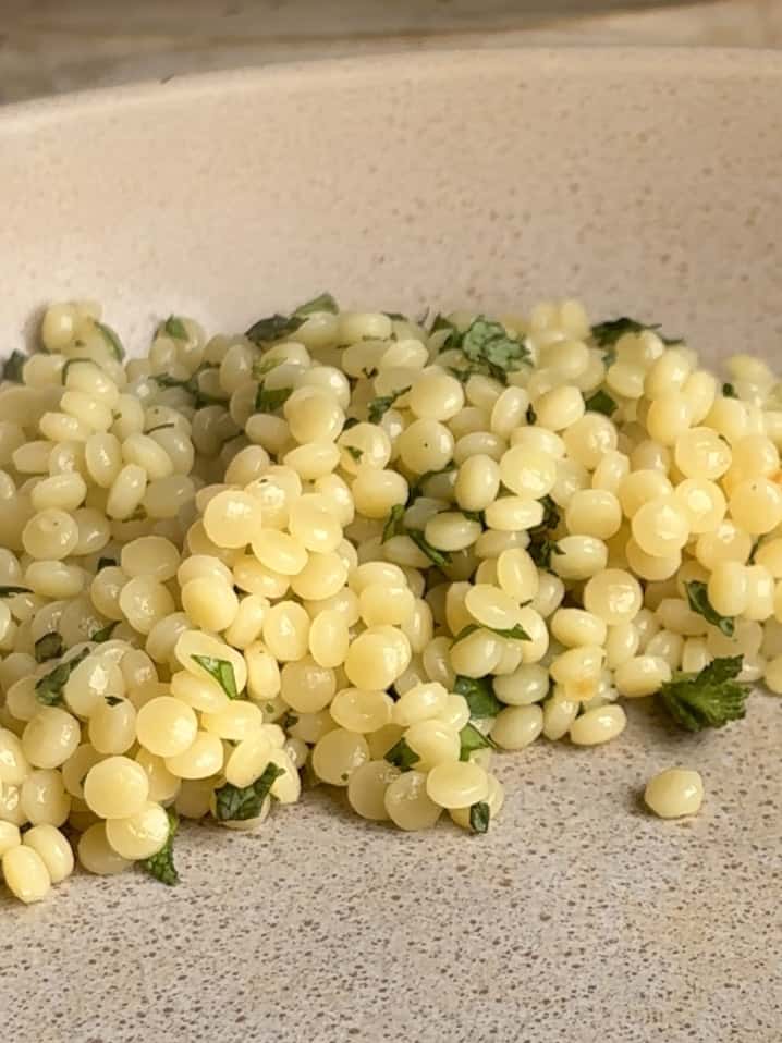 a sideview shot of the cooked pearl couscous scattered with finely chopped parsley and mint in the serving dish.