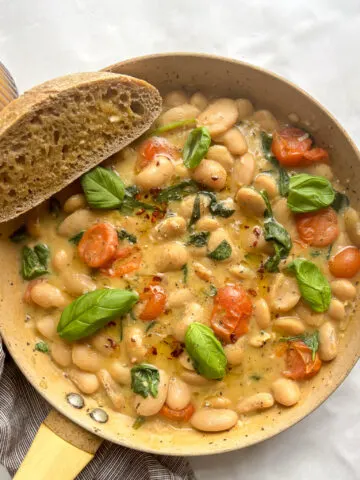 overhead shot of a pan of creamy butter beams topped with basil and a slice of crusty bread on the side