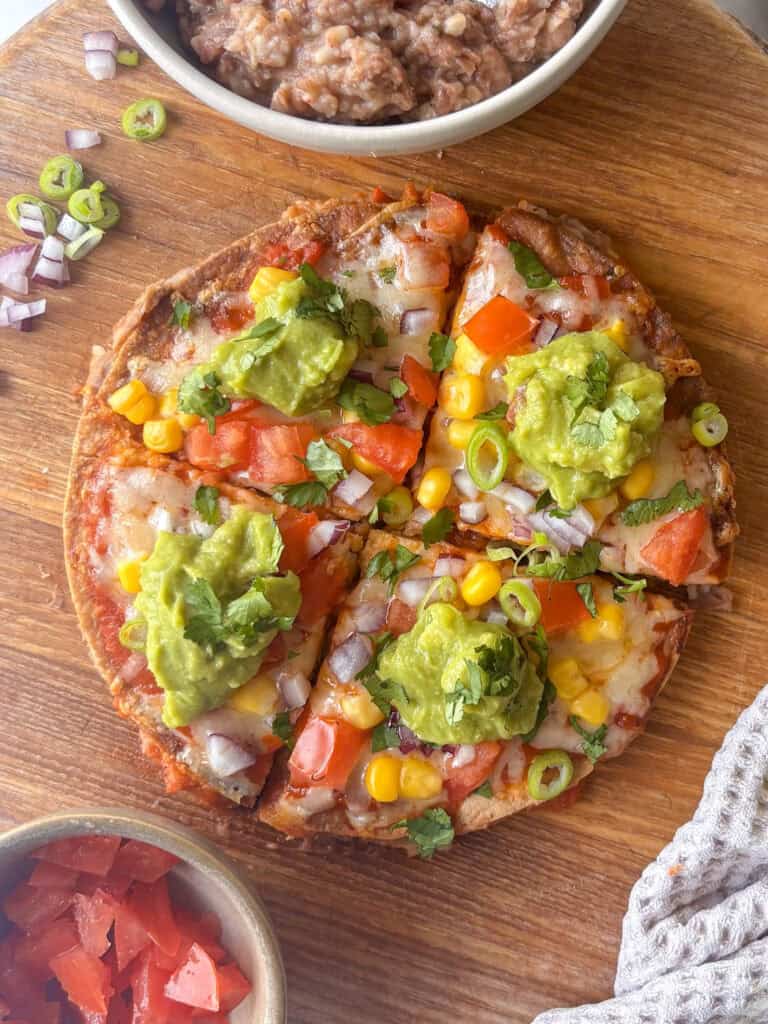 mexican pizza on a chopping board, topped with vegetables and a spoonful of guacamole on each quarter
