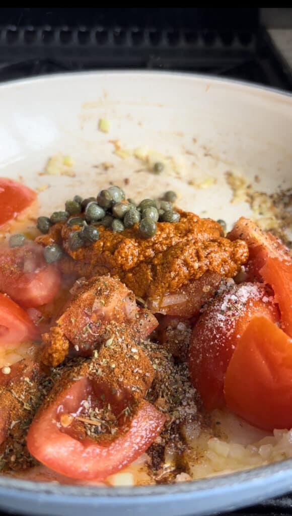 fresh tomatoes, red poesto, capers, and seasonings are added to the casserole dish with the onion and garlic