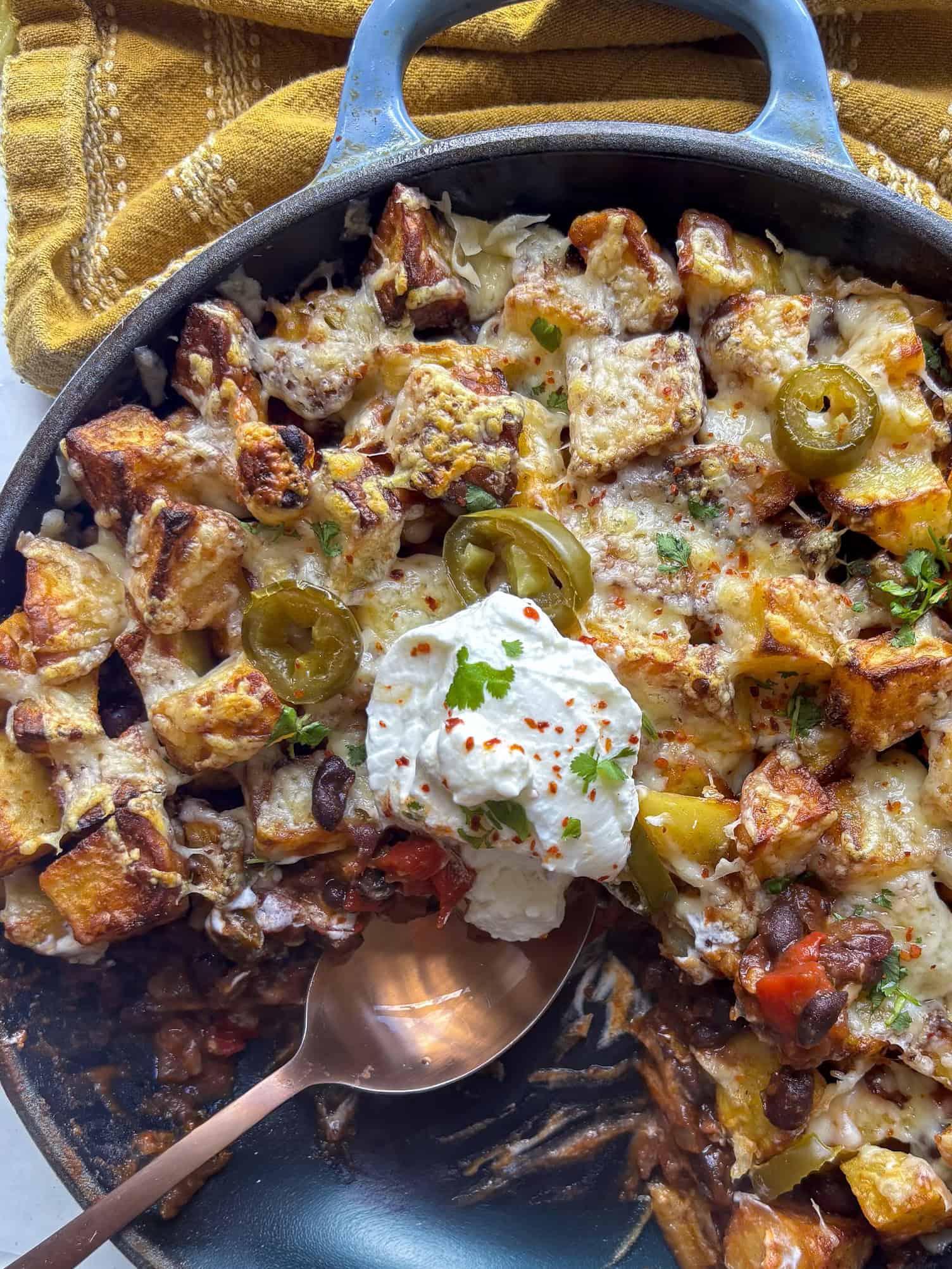 a cast iron skillet of smoky black beans topped with crispy cubed potatoes and melted cheese, jalapenos, and a spoonful of yogurt.