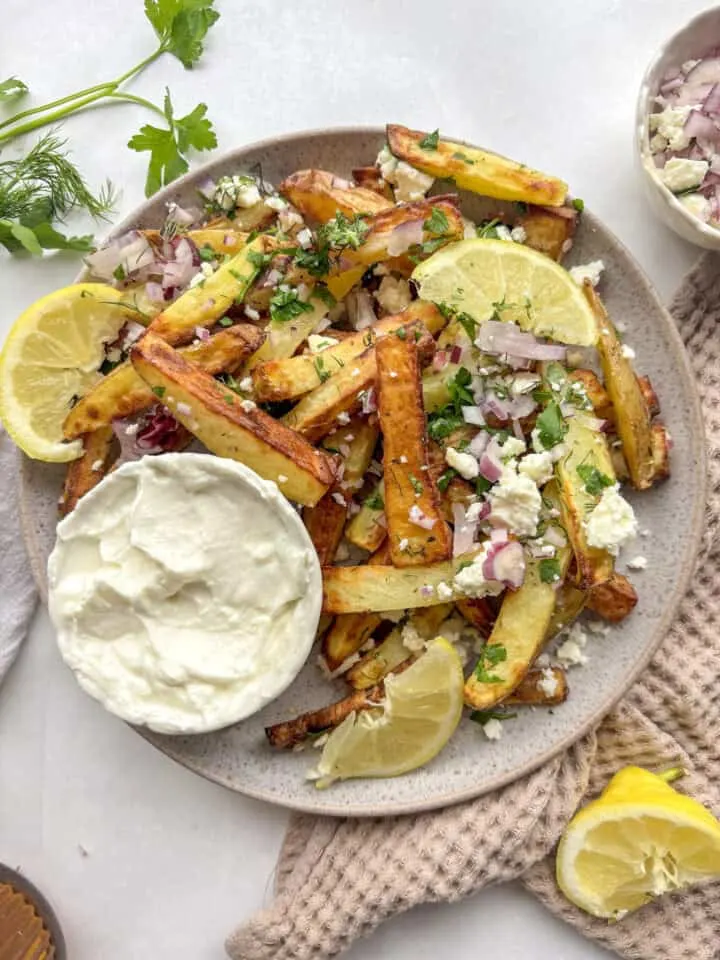 Crispy golden potato wedges topped with diced red onion, crumbled feta, cilantro, lime wedges, and sour cream dip on gray ...