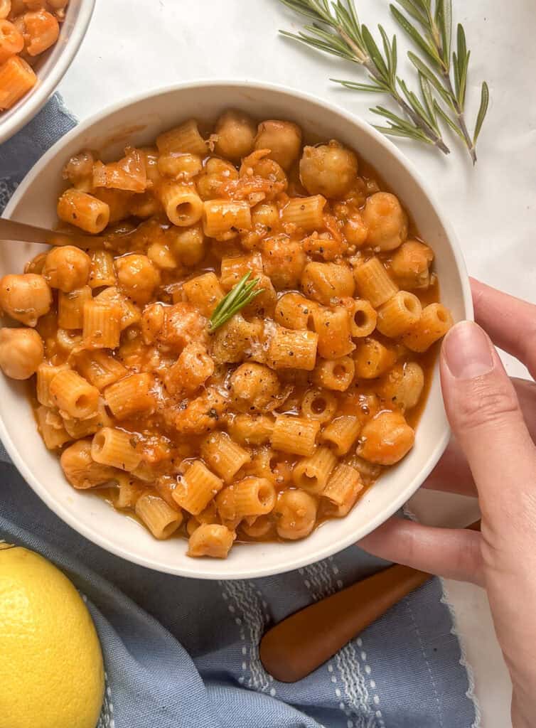 a white bowl of pasta and chickpeas, topped with black pepper, hard cheese, and olive oil