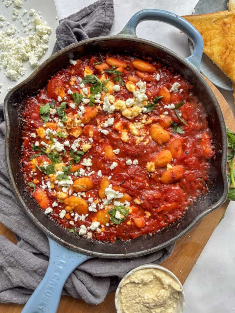 an overhead shot of a skillet of gigantes plaki scattered with feta and parsley, surrounded by hummus and crusty bread