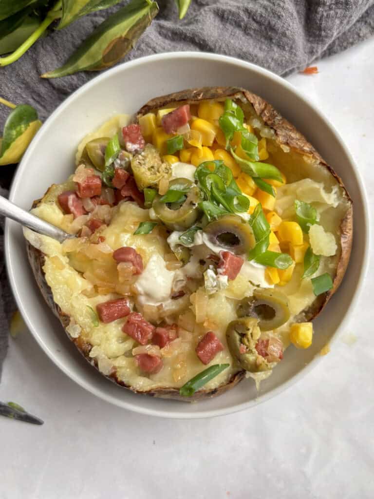 a bowl of kumpir (stuffed baked potato) loaded with sour cream, vegetarian bacon and onions, chopped olives, sweetcorn, and chives