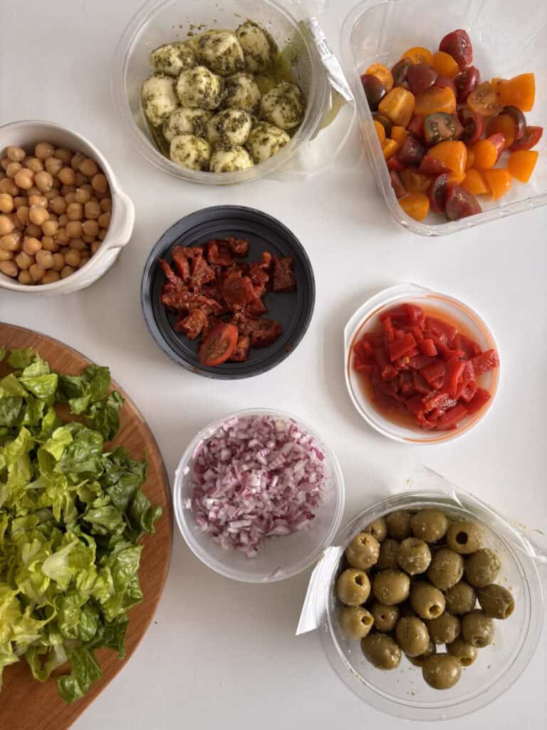 ingredients for italian chopped salad laid out ready to be assembled: olives, red pepper, basil, red onion, chickpeas, lettuce, hard cheese, tomatoes, sun-dried tomatoes, and mozzarella pearls