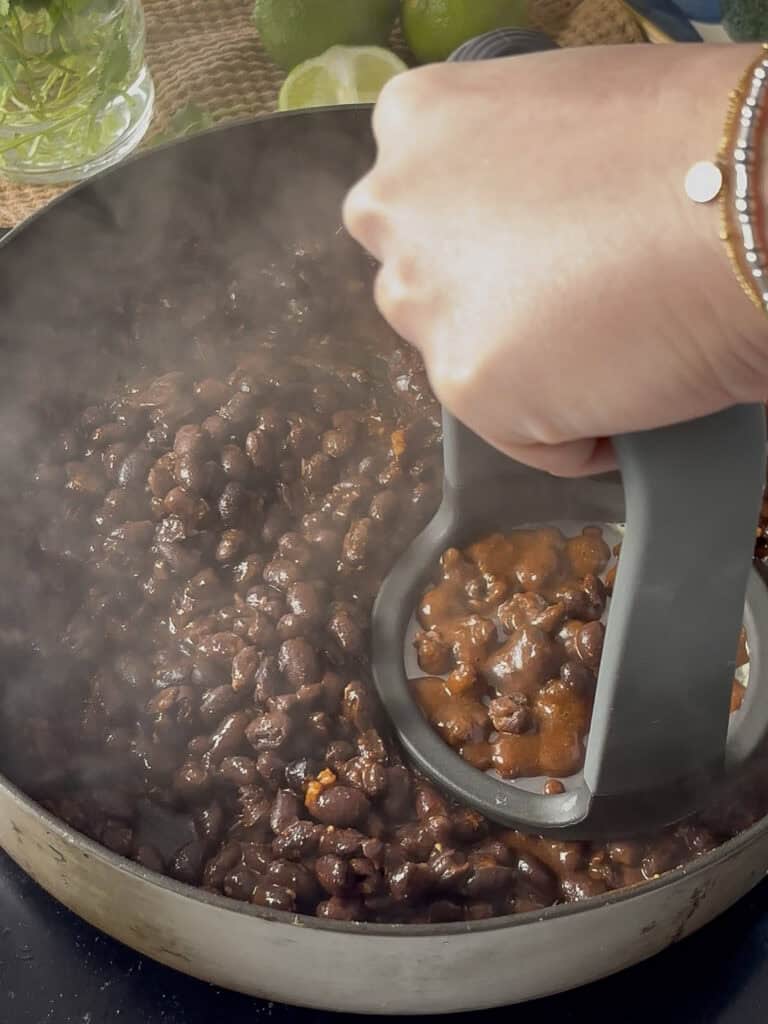 the black bean mixture in a saucepan with a potato masher being used to mash the beans