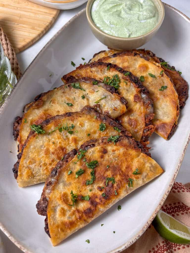 a plate of crispy black bean tacos with fresh cilantro scattered on top and a dipping bowl of creamy lime and cilantro sauce