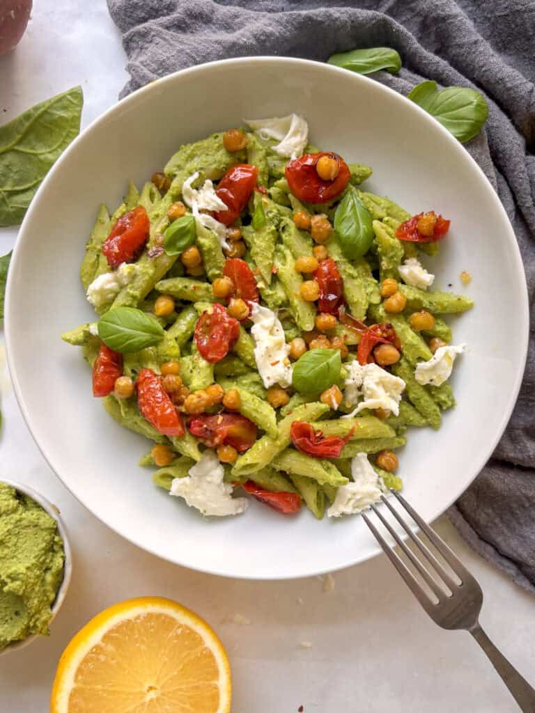a white bowl of chickpea pesto pasta topped with roasted chickpeas and cherry tomatoes, as well as mozzarella and basil leaves