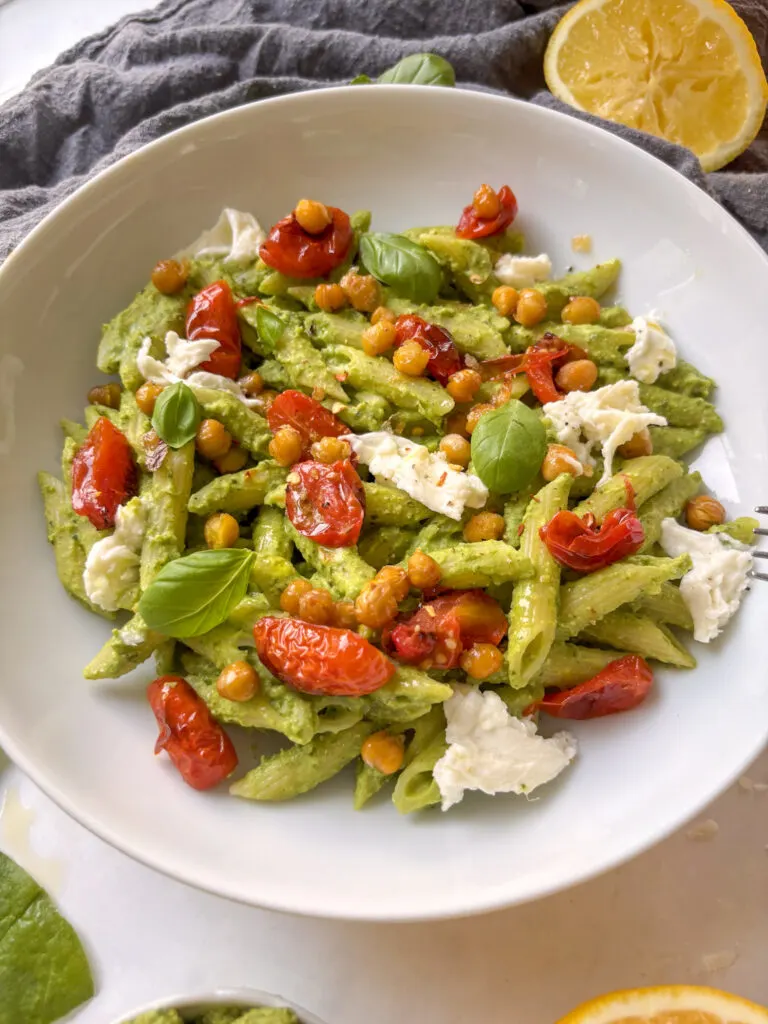 a white bowl of chickpea pesto pasta topped with roasted chickpeas and cherry tomatoes, as well as mozzarella and basil leaves