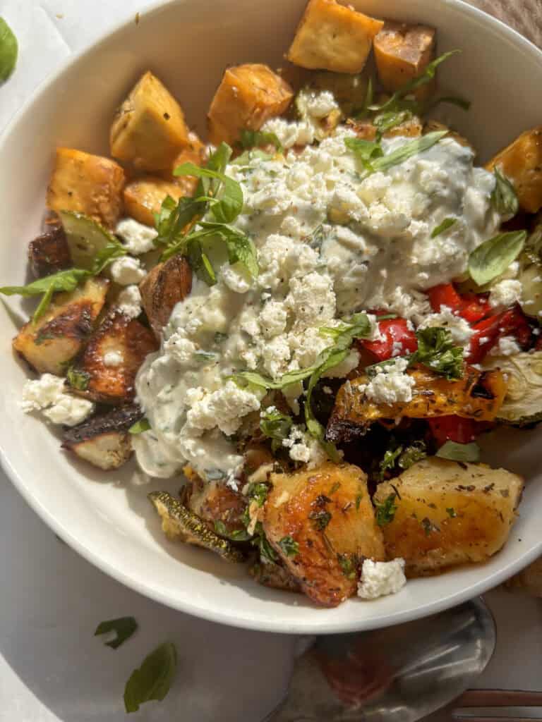 a bowl of baked tofu, potatoes, and vegetables topped with cucumber yogurt dip and tzatziki