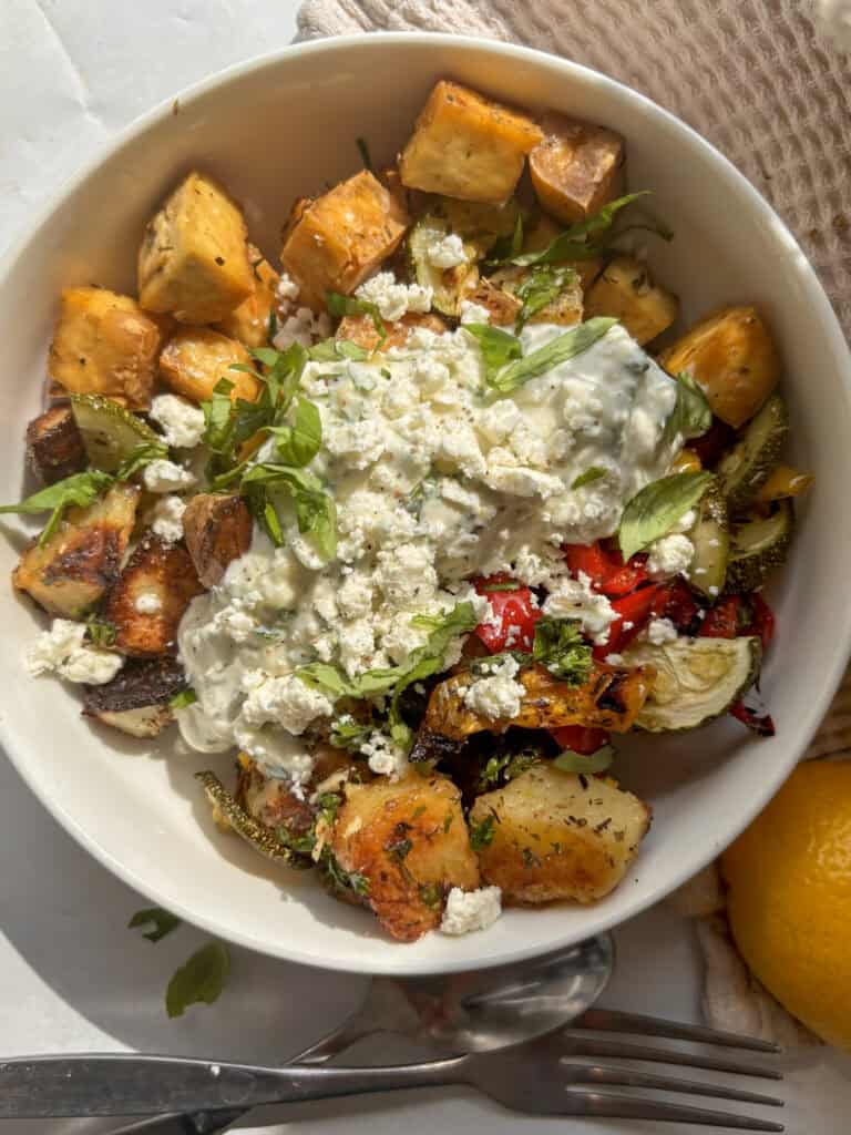 a white bowl with baked tofu, potatoes, and Mediterranean vegetables topped with tzatziki, feta, and basil