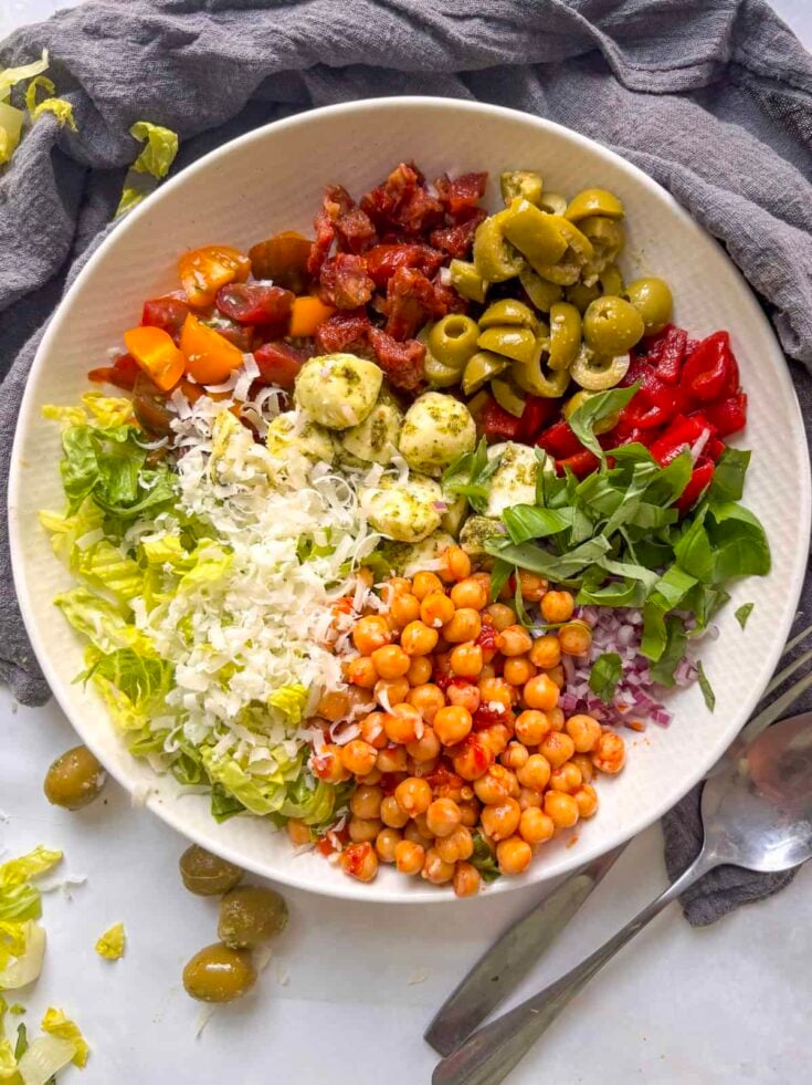 a italian chopped salad assembled before mixing, with olives, red pepper, basil, red onion, chickpeas, lettuce, hard cheese, tomatoes, sun-dried tomatoes, and mozzarella pearls