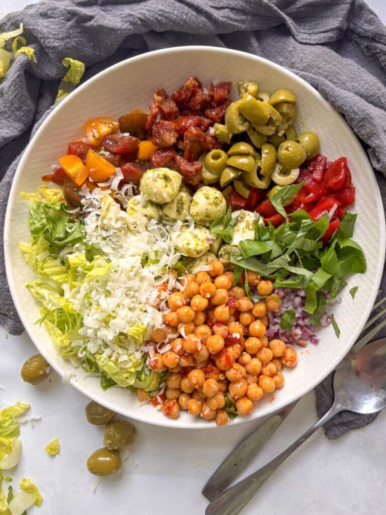 a italian chopped salad assembled before mixing, with olives, red pepper, basil, red onion, chickpeas, lettuce, hard cheese, tomatoes, sun-dried tomatoes, and mozzarella pearls