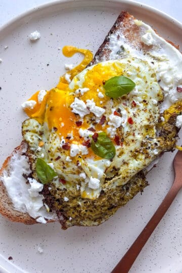 pesto fried egg laid on top of sourdough toast with a garlic yoghurt base. The egg is topped with crumbled feta and red chili flakes